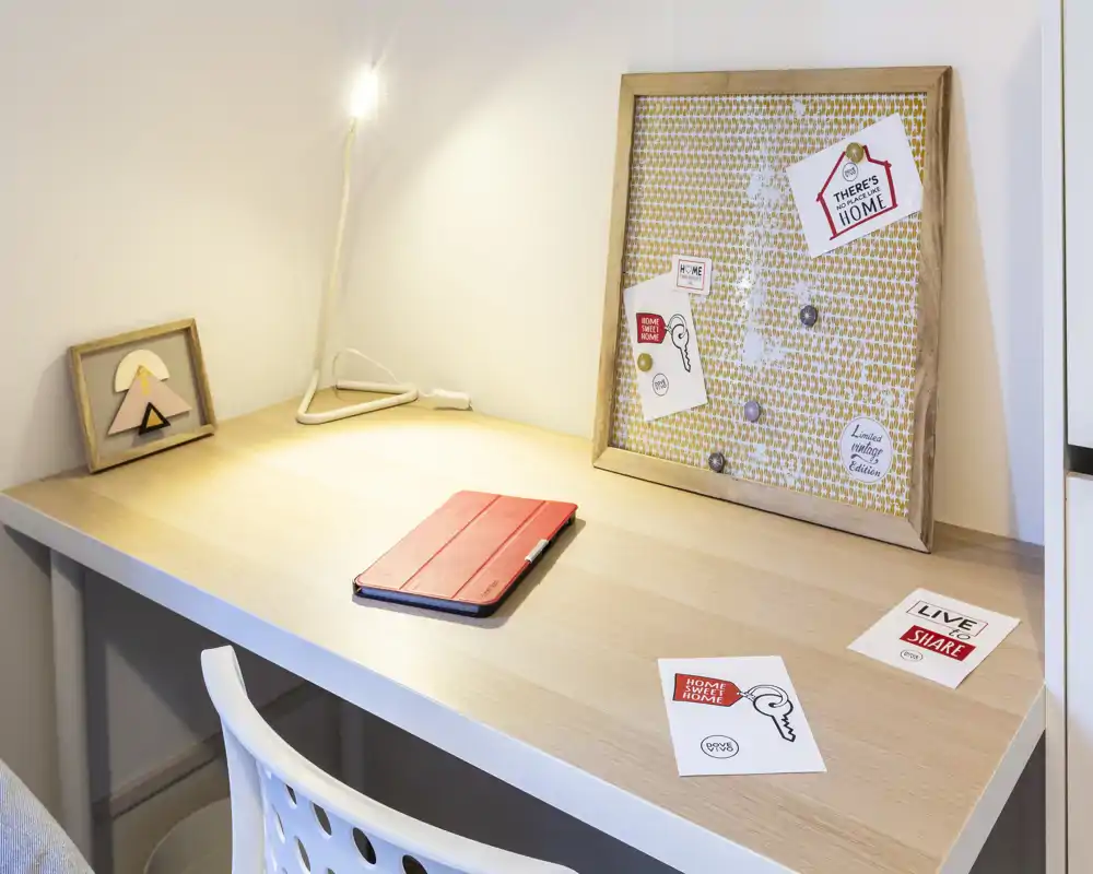 Bright and tidy workspace corner with a wooden desk, a small lamp, decorative pinboard and a tablet — ideal for focused work or study.