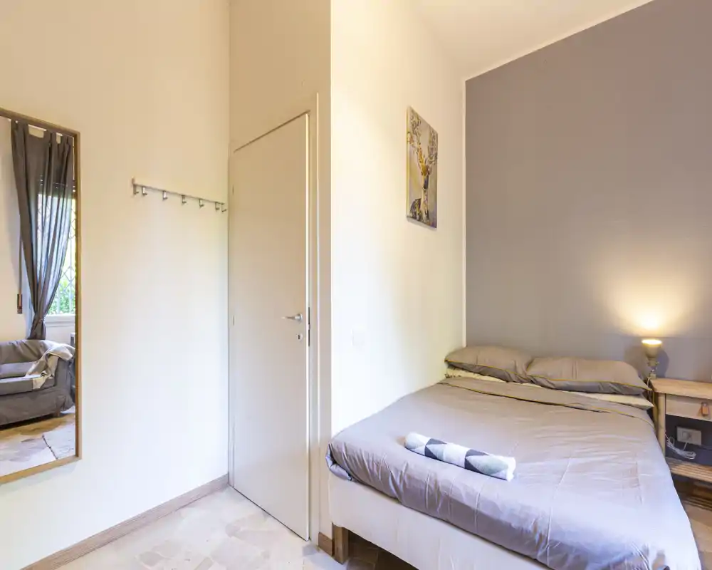 Comfortable double bedroom with neutral linens, bedside table and warm lamp. Natural light from the window and a decorative wall painting create a welcoming, tidy space ideal for short stays.