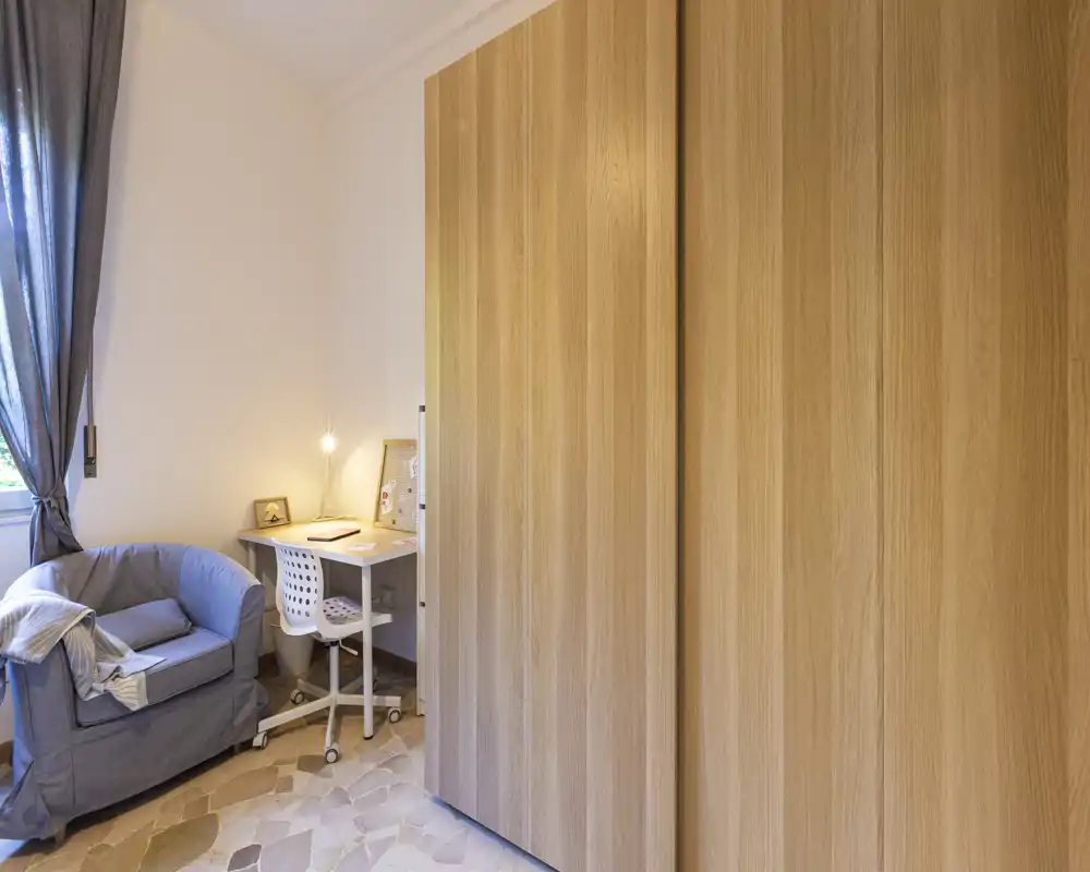 Small bedroom corner with a large wooden wardrobe, a cozy armchair and a compact desk with task lamp — bright and tidy, suitable for working or relaxing.