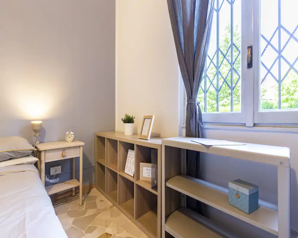 Bright, cozy single bedroom corner with a bedside table, lamp and shelving under a large window providing natural light.