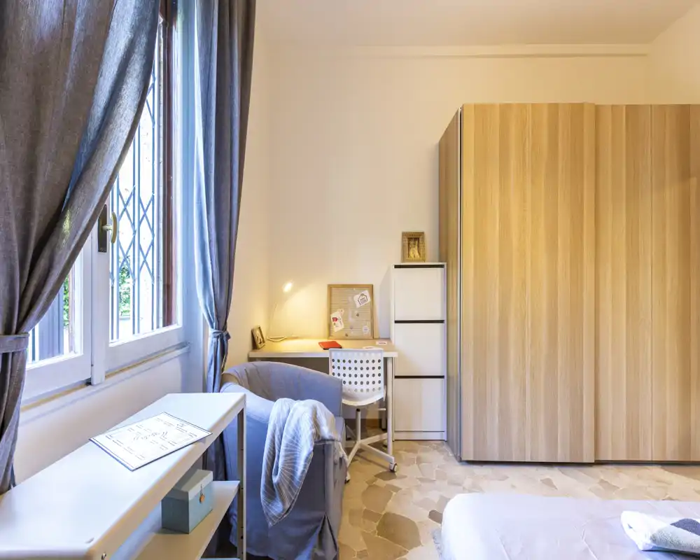 Bright, well-styled bedroom corner with a visible bed, wooden wardrobe, desk with task lamp and a cozy armchair by the window.