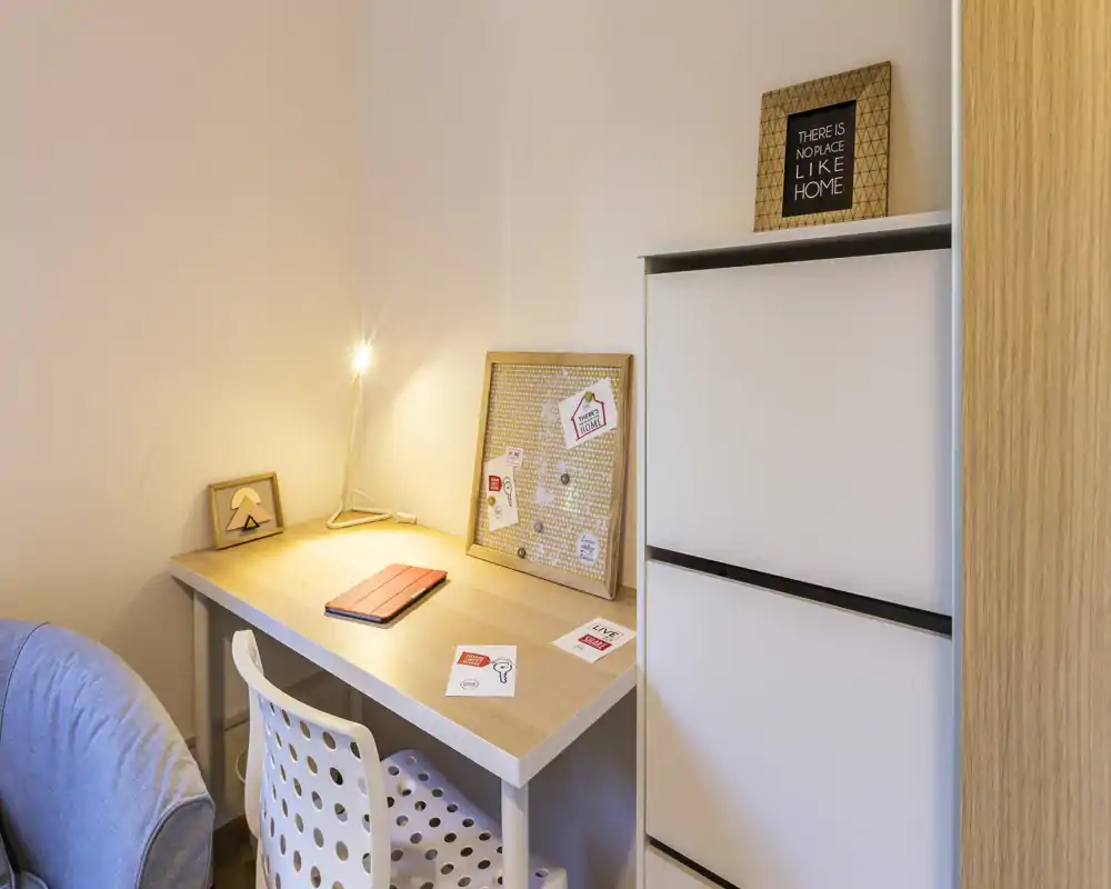 A cozy bedroom workspace with a wooden desk, perforated chair and warm desk lamp; decorative notes and a small shelving unit create a tidy, inviting corner.