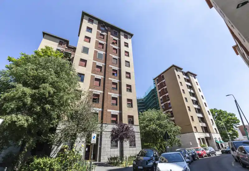 Street-level view of an apartment building façade with parked cars and trees under a clear blue sky, suitable for showing the building exterior and neighborhood.