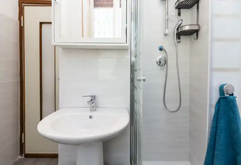 Compact, modern bathroom with a pedestal sink, mirrored cabinet and a glass-enclosed shower with handheld showerhead. Bright, clean finishes and a turquoise towel add a fresh touch.