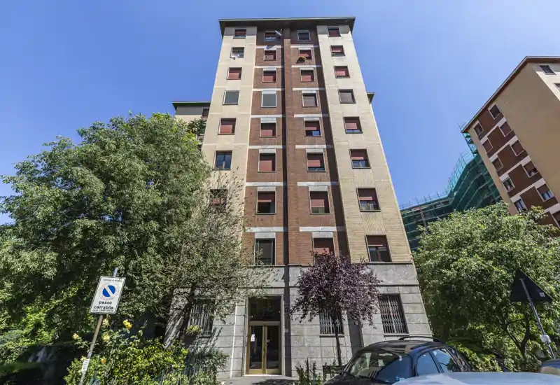 Exterior view of a multi-story residential building with trees, clear blue sky and a visible entrance — suitable for showing the property façade and street presence.
