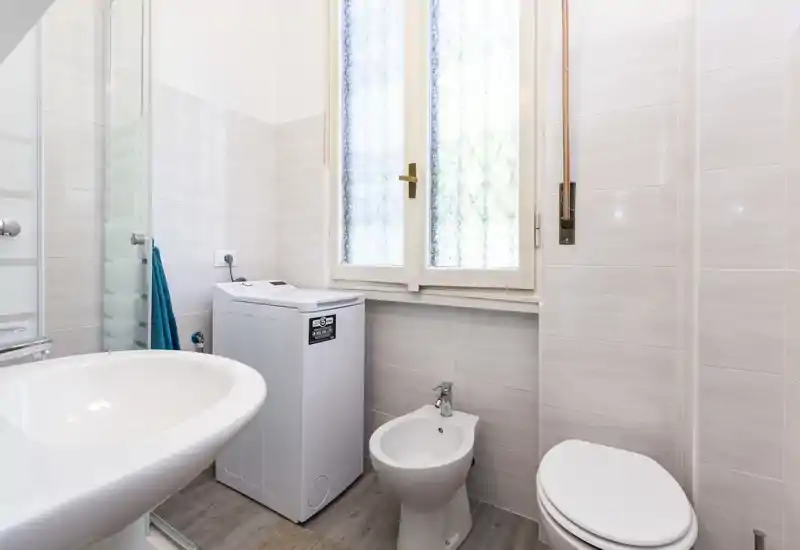 Bright, well-lit bathroom with a sink, bidet, toilet and a compact washing machine under a frosted window; neutral tiles and wood-effect floor create a clean, modern look.