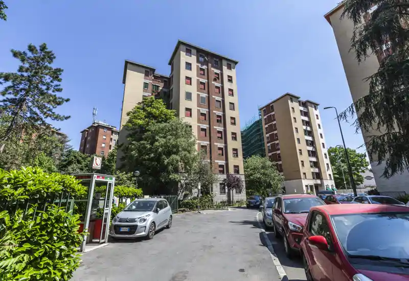 Exterior view of residential apartment buildings with a paved parking area and parked cars under a clear blue sky.