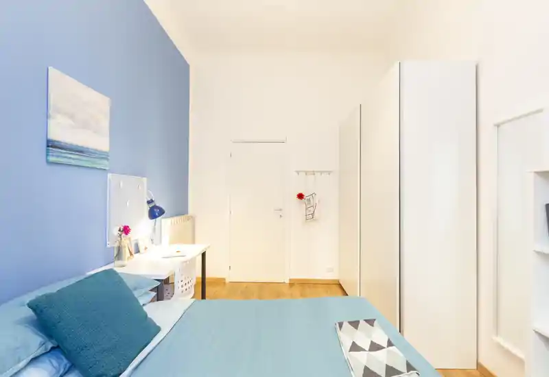 Camera da letto moderna e luminosa con biancheria turchese, scrivania e grande armadio bianco. La stanza è ben illuminata e arredata con gusto con cuscini e quadri.