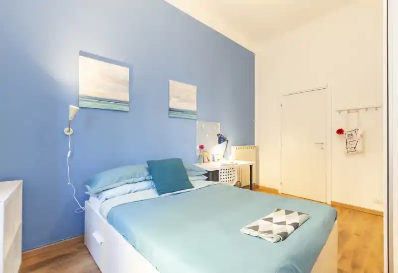 Camera da letto luminosa e ben arredata con letto matrimoniale davanti a una parete blu, lampade e una scrivania: perfetta per un soggiorno confortevole.