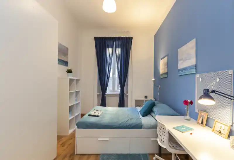 Camera da letto luminosa e ben arredata con letto matrimoniale, arredi coordinati blu, scrivania e scaffalatura — perfetta per un soggiorno confortevole.