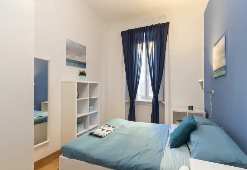 Camera da letto luminosa e ordinata con letto matrimoniale in biancheria turchese, cuscini morbidi e finestra con tende blu scuro che garantiscono luce naturale.