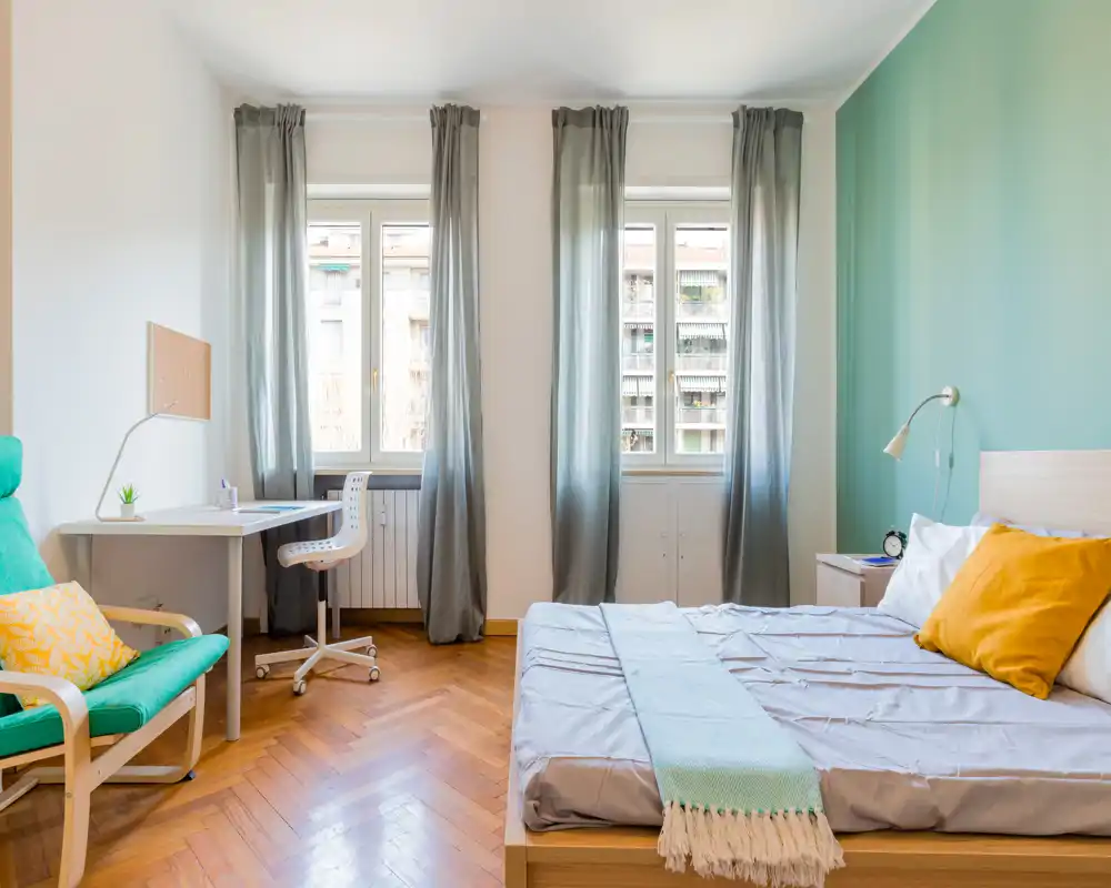 Camera da letto luminosa e arredata con un comodo letto matrimoniale, comodino e una postazione di lavoro vicino alla finestra; parete accento turchese e pavimento in legno creano un'atmosfera accogliente e moderna.