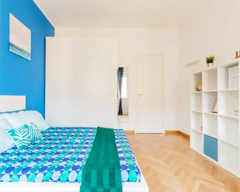 Camera da letto moderna e luminosa con letto matrimoniale con biancheria geometrica colorata, parete accentuata blu intenso, armadio bianco e scaffalatura, e pavimento in parquet caldo.