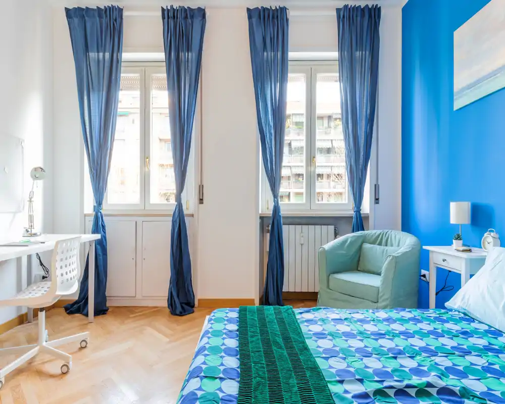 Camera da letto luminosa e ben arredata con letto matrimoniale a fantasia, poltrona accogliente e luce naturale da due finestre alte — perfetta per un annuncio di affitto.