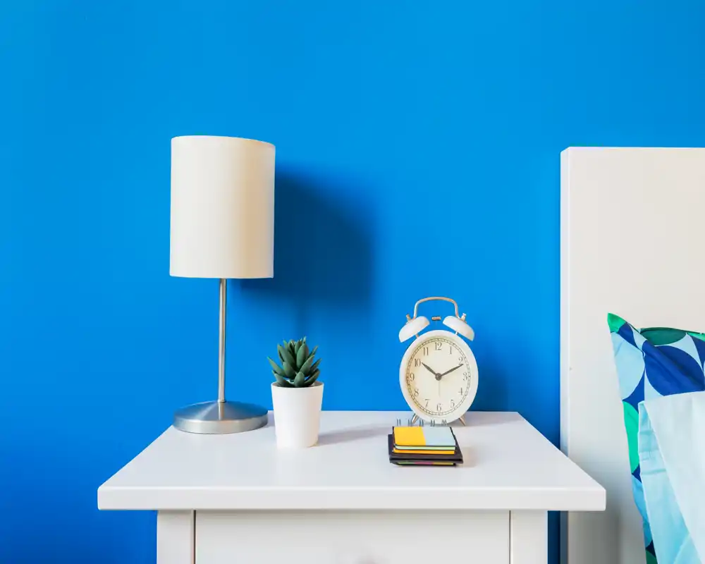 Scena luminosa e moderna del comodino in camera da letto: comodino bianco su parete blu brillante, lampada, sveglia e cuscino decorativo sul bordo del letto.