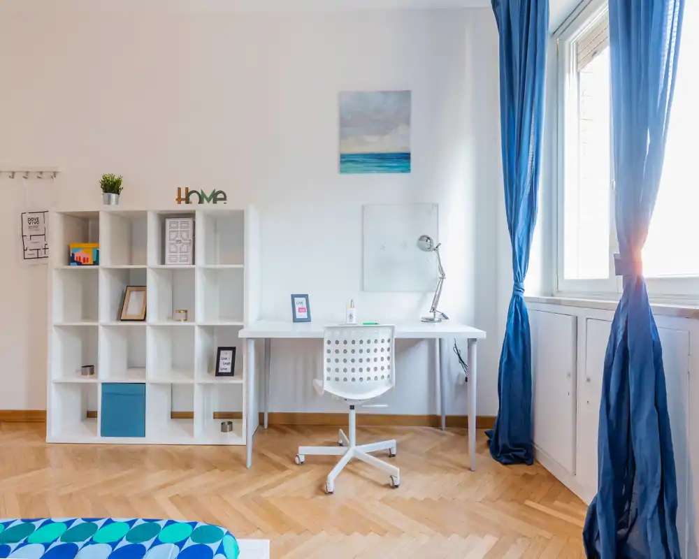 Camera da letto luminosa con angolo studio minimale: scrivania bianca e sedia accanto alla finestra con tende blu, una libreria modulare per lo stoccaggio e un angolo letto visibile con copriletto blu a fantasia.
