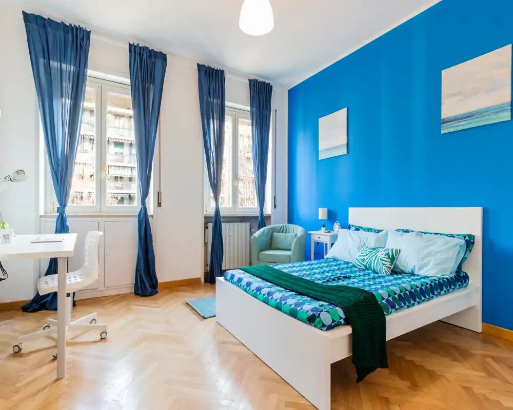 Camera luminosa e moderna con letto bianco su una parete blu acceso, biancheria a motivi, una poltrona accogliente e una postazione di lavoro vicino a tre ampie finestre che illuminano la stanza con luce naturale.