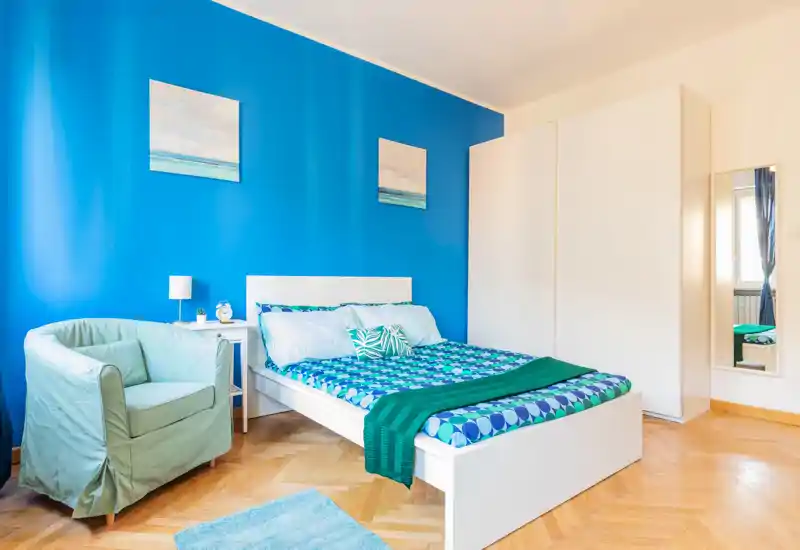 Camera da letto luminosa e ben arredata con letto bianco su parete accentuata blu, biancheria a fantasia e una poltrona accogliente — perfetta per un annuncio moderno.