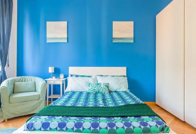 Camera da letto luminosa e moderna con letto matrimoniale dal copriletto decorato su una parete blu acceso, comodino con lampada e una comoda poltrona — perfetta per un soggiorno vivace.