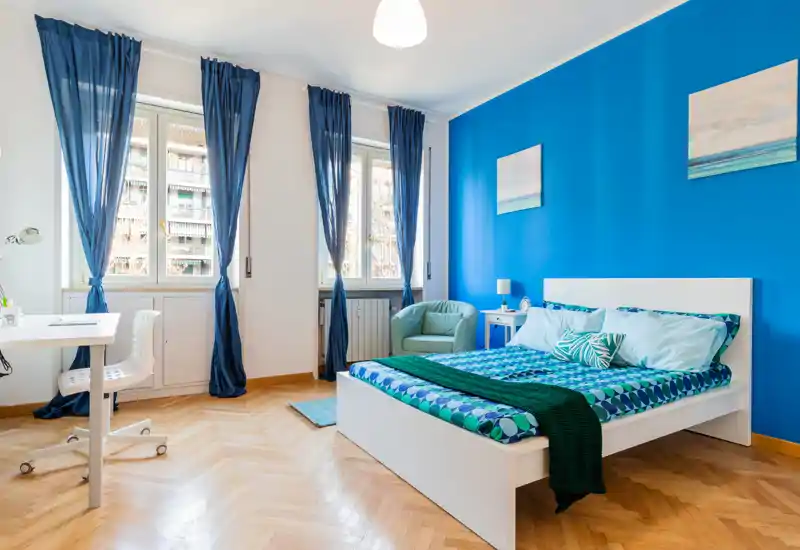 Camera luminosa e moderna con letto bianco su una parete blu acceso, biancheria a motivi, una poltrona accogliente e una postazione di lavoro vicino a tre ampie finestre che illuminano la stanza con luce naturale.