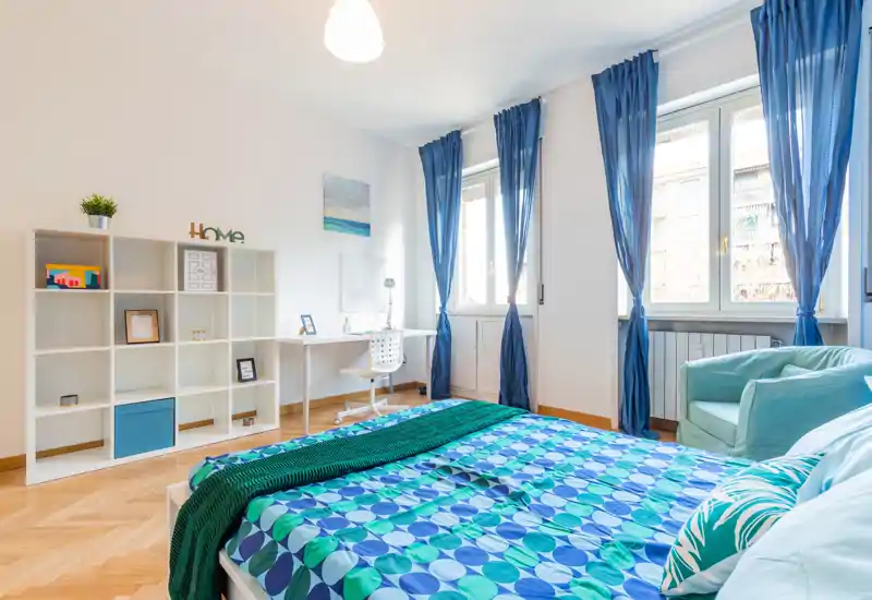 Camera da letto luminosa e ben arredata con letto matrimoniale con biancheria colorata, grandi finestre con tende blu, una scrivania vicino alla finestra e scaffalature per riporre oggetti — uno spazio arioso e confortevole ideale per rilassarsi o lavorare.