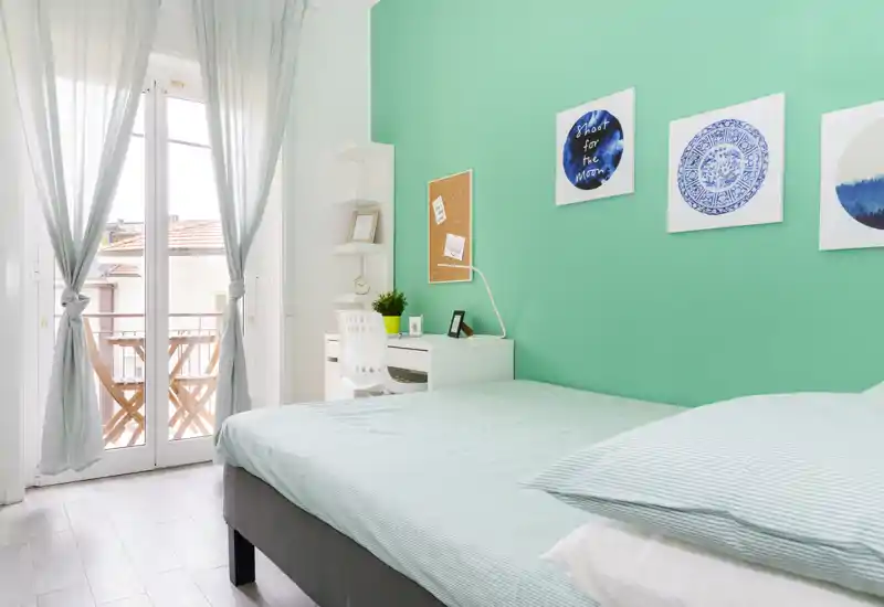 Chambre moderne et lumineuse avec lit double confortable, mur accent vert menthe, petit bureau et balcon apportant une belle lumière naturelle.