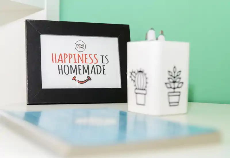 Gros plan décoratif montrant un cadre avec l'inscription « Happiness is homemade » et un petit pot blanc avec des illustrations de plantes sur un fond turquoise. L'image met l'accent sur des accessoires de table plutôt que sur une pièce identifiable.