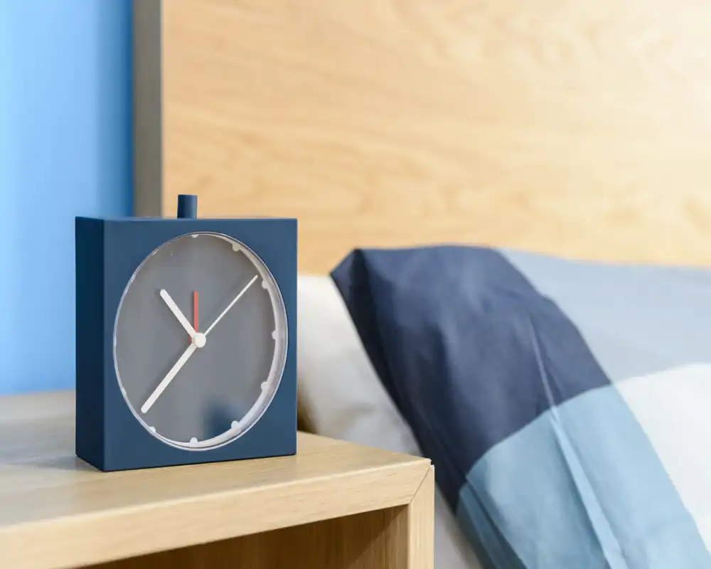 Gros plan d'un coin de chevet montrant un réveil bleu moderne sur une table de nuit en bois et une partie de l'oreiller et de la literie aux tons bleus.