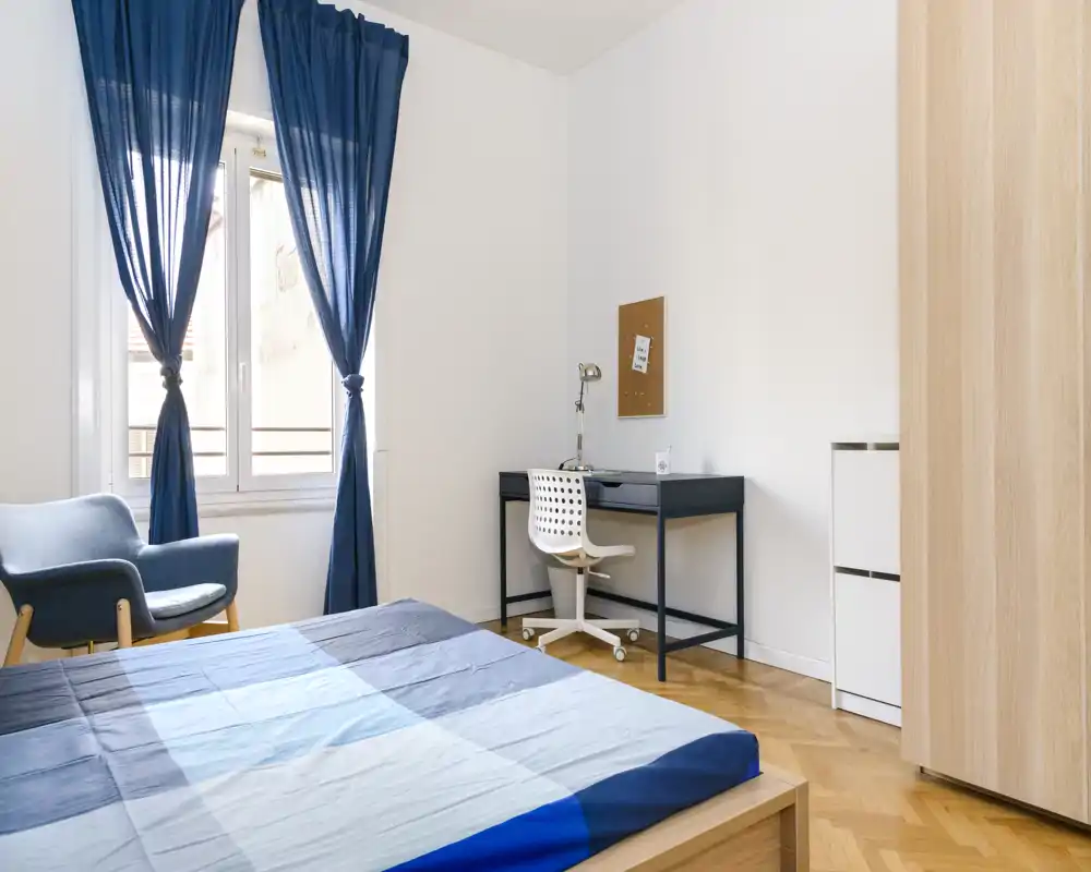 Chambre lumineuse et moderne avec lit en bois et linge de lit bleu, fenêtre apportant une belle lumière naturelle; bureau et fauteuil confortable inclus — idéale pour le repos et l'étude.