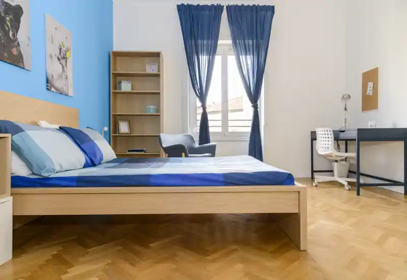 Chambre lumineuse et moderne avec lit en bois, linge de lit bleu et rideaux assortis. La pièce comprend un bureau, une étagère et un fauteuil près de la fenêtre, idéale pour se reposer et travailler.