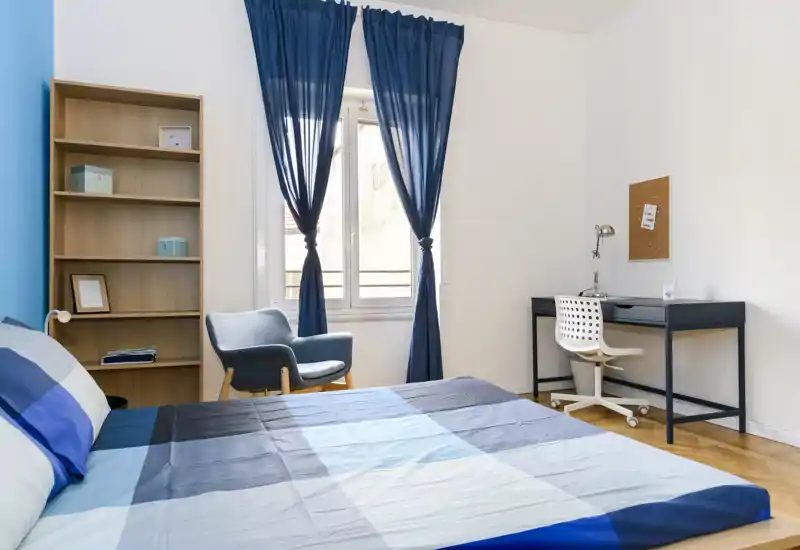 Chambre moderne et lumineuse avec un grand lit bleu et gris, un fauteuil près de la fenêtre, étagères et bureau minimaliste — idéale pour étudiants ou courts séjours.