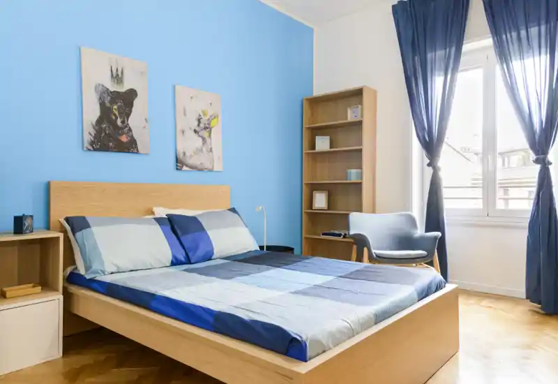 Chambre moderne et lumineuse avec un lit en bois, linge de lit bleu coordonné et une grande fenêtre apportant une belle lumière naturelle.