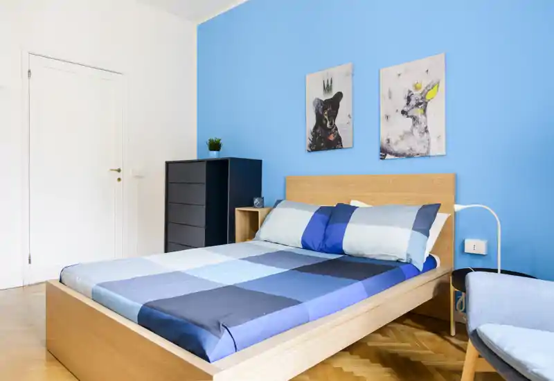 Chambre moderne et lumineuse avec un lit double en bois habillé de linge de lit bleu à motifs géométriques, oreillers assortis, lampe de chevet et tableaux décoratifs sur un mur bleu.