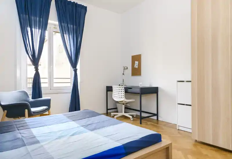 Chambre lumineuse et moderne avec lit en bois et linge de lit bleu, fenêtre apportant une belle lumière naturelle; bureau et fauteuil confortable inclus — idéale pour le repos et l'étude.