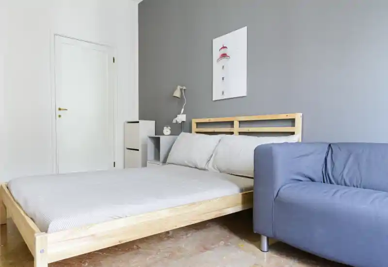 Chambre lumineuse et minimaliste avec un lit double en bois, linge de lit neutre et un petit canapé à côté. La pièce présente un mur gris, une étagère de chevet avec lampe et une affiche simple, pour une ambiance moderne et apaisante.