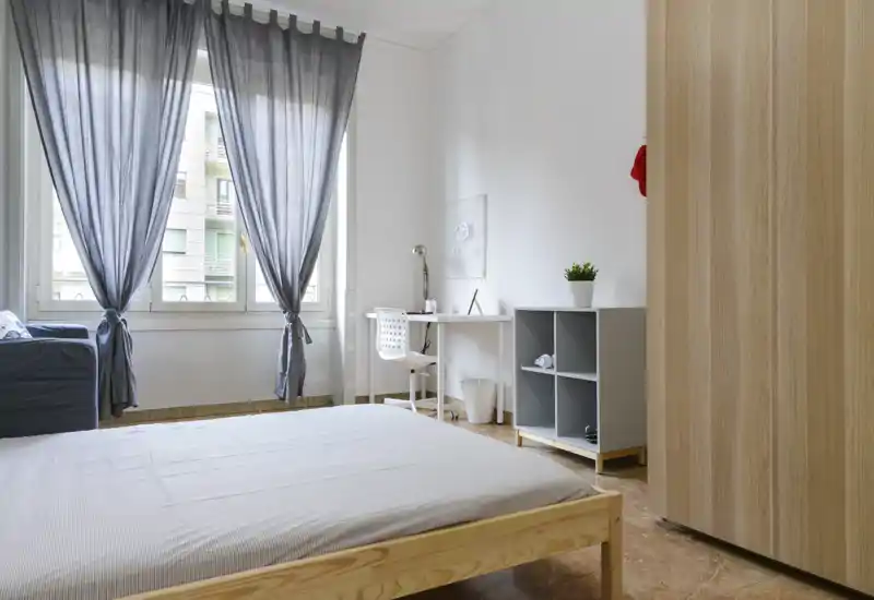 Chambre lumineuse et minimaliste avec lit sur cadre en bois, grande fenêtre aux rideaux gris, petit bureau et étagère cubique — parfaite pour une personne ou de courts séjours.