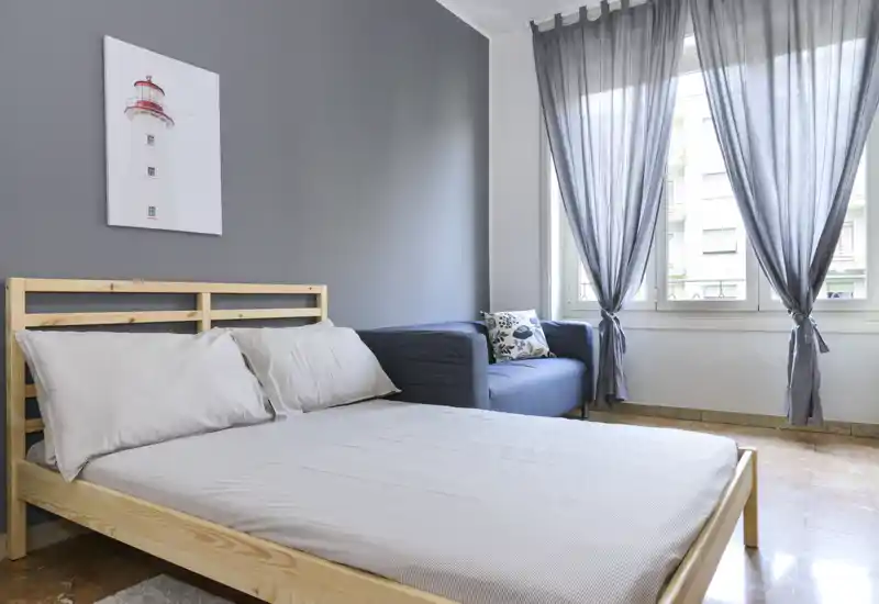 Chambre lumineuse au style épuré avec un lit à cadre en bois et linge de lit clair, un petit canapé près de la fenêtre et une douce lumière naturelle filtrée par des voilages.