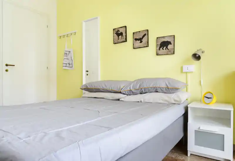 Chambre lumineuse et moderne avec un lit double bien fait, linge de lit gris, une table de chevet avec lampe et horloge, et des cadres muraux discrets sur un mur jaune pâle.