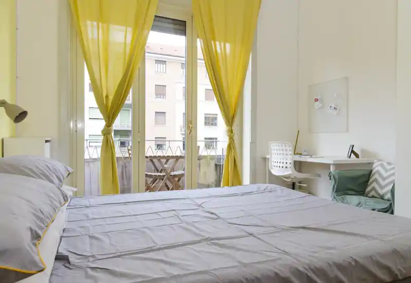 Chambre lumineuse et moderne avec grand lit donnant sur un balcon encadré de rideaux jaunes, un petit bureau et un fauteuil confortable — idéale pour un séjour reposant.