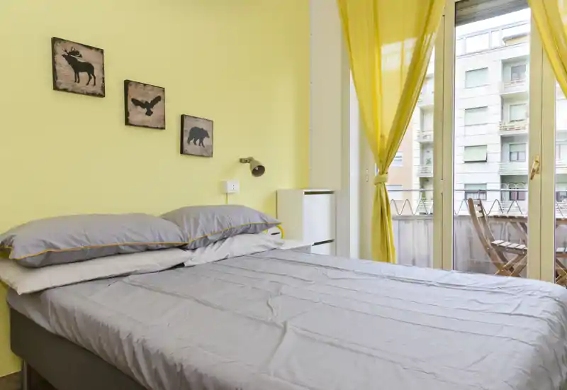 Chambre lumineuse et soignée avec un lit double, linge de lit gris et des murs jaune. Grandes portes vitrées s'ouvrent sur un petit balcon et apportent une belle lumière naturelle.