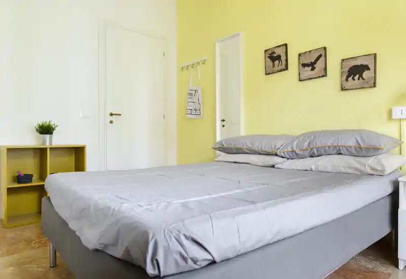 Chambre lumineuse et bien rangée avec un grand lit, linge de lit gris et un mur jaune chaleureux. Décoration simple et belle lumière naturelle pour une ambiance accueillante et fraîche, idéale pour une annonce locative.