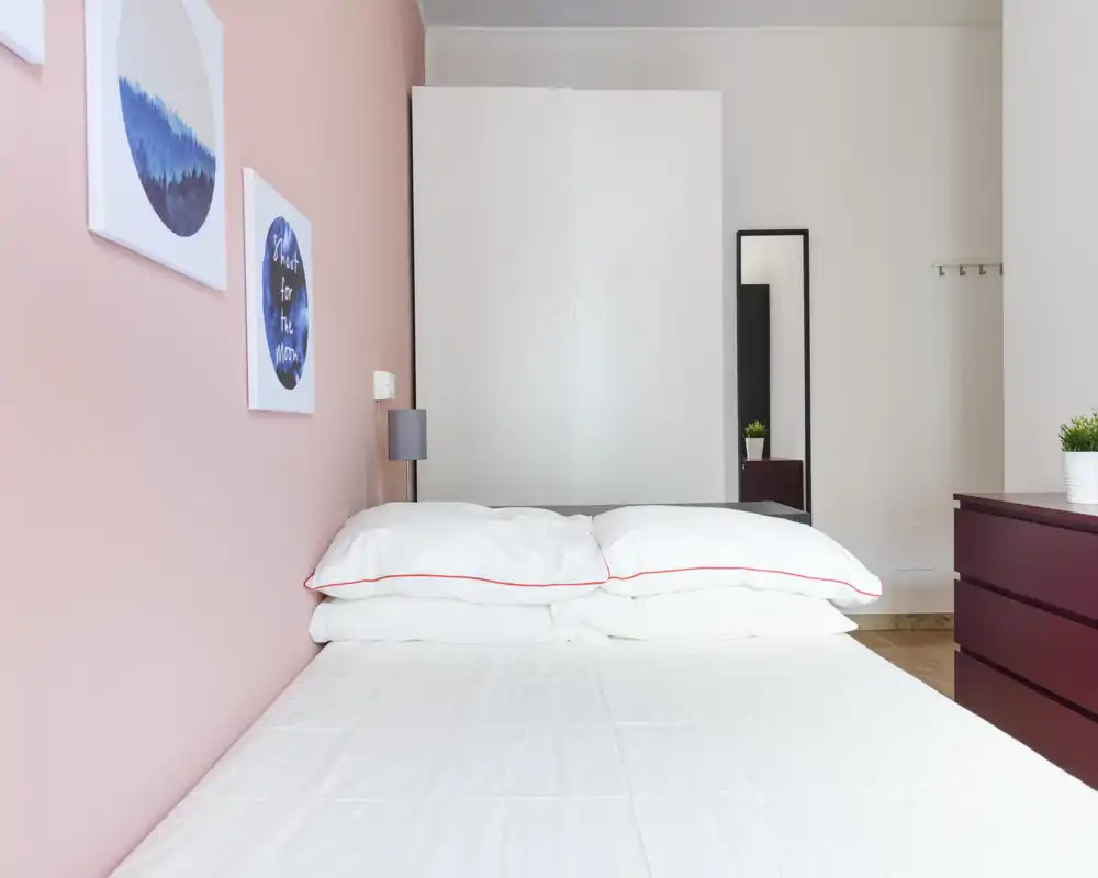 Dormitorio moderno y luminoso con cama doble bien hecha, ropa de cama blanca y pared rosa; armario y espejo al fondo que aportan almacenamiento.
