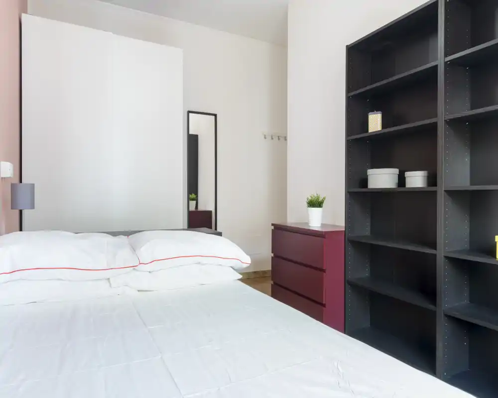 Dormitorio moderno y luminoso con una cama doble bien hecha en primer plano, ropa de cama blanca con ribete rojo, un espejo alto y estanterías abiertas a la derecha.