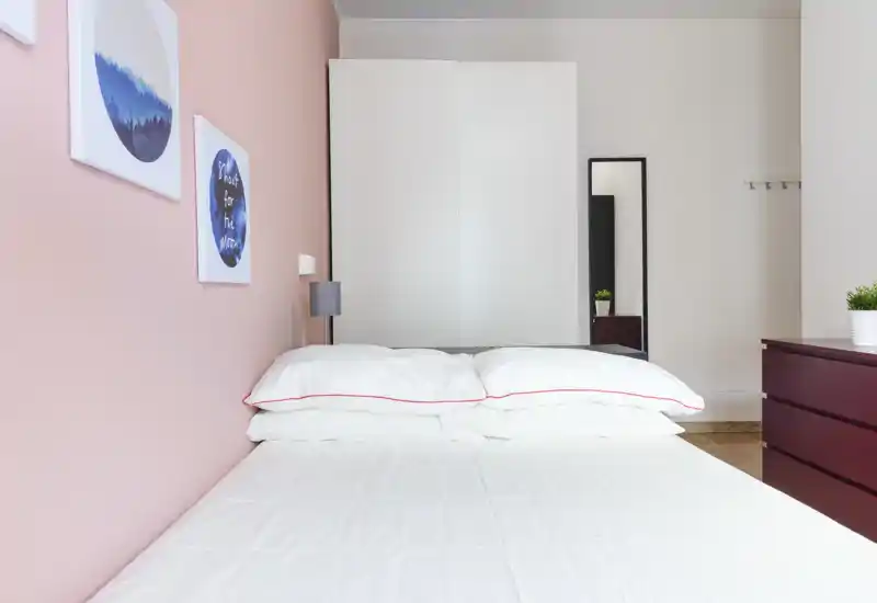 Dormitorio moderno y luminoso con cama doble bien hecha, ropa de cama blanca y pared rosa; armario y espejo al fondo que aportan almacenamiento.