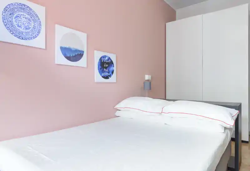 Dormitorio moderno y luminoso con cama doble bien hecha, pared rosa suave y armario minimalista — líneas limpias y decoración relajante, perfecto para anuncios de alquiler.