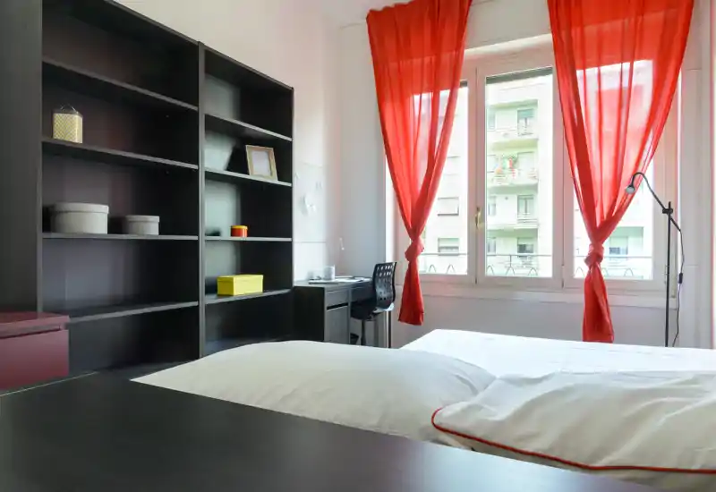 Dormitorio luminoso y bien arreglado con una gran cama en primer plano, estantería y escritorio junto a la ventana con cortinas rojas llamativas.