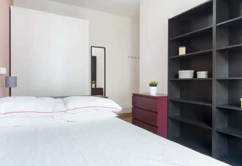 Dormitorio moderno y luminoso con una cama doble bien hecha en primer plano, ropa de cama blanca con ribete rojo, un espejo alto y estanterías abiertas a la derecha.
