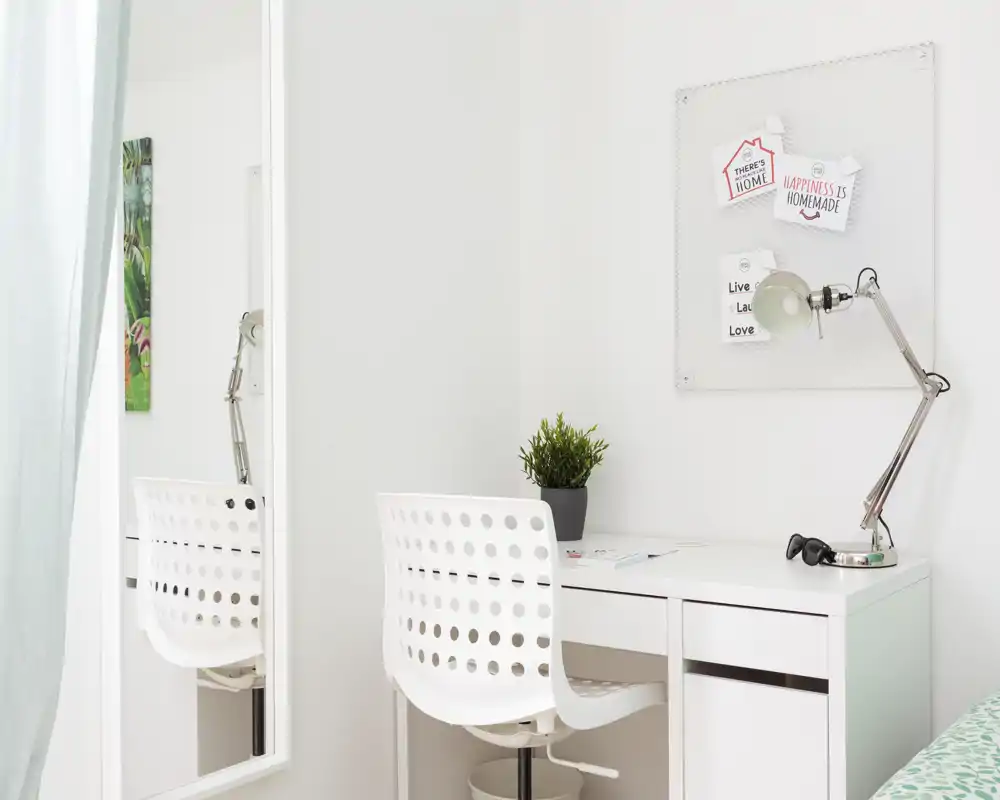 Bright, minimal bedroom corner with a white desk and perforated chair, a bedside/desk lamp and a small plant; soft natural light and clean, modern styling.