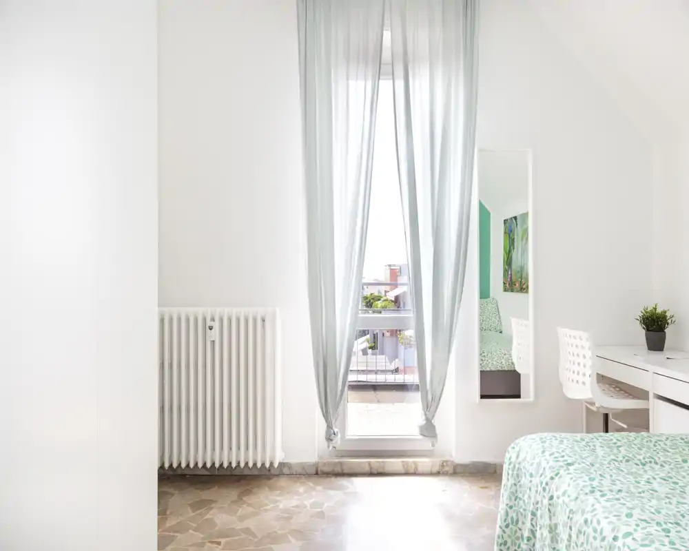 Bright, minimal bedroom with a patterned bedspread, a desk and a window that opens to a small balcony — clean white walls and soft natural light create a calm atmosphere.