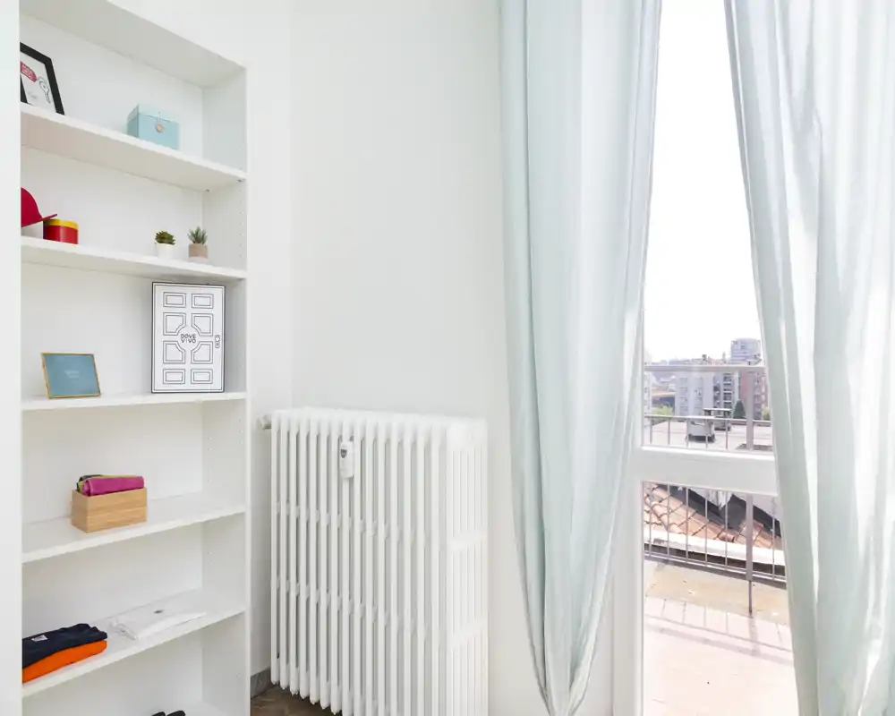 Bright interior corner with shelving and a radiator opening onto a small balcony view — soft curtains frame a sunlit city terrace visible through the window.